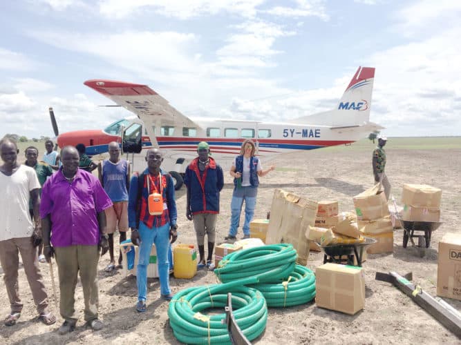 Kuvassa Etelä-Sudanilaisia miehiä seisomassa MAF:n koneen edessä vesiputkien ja pahvilaatikoiden kanssa.